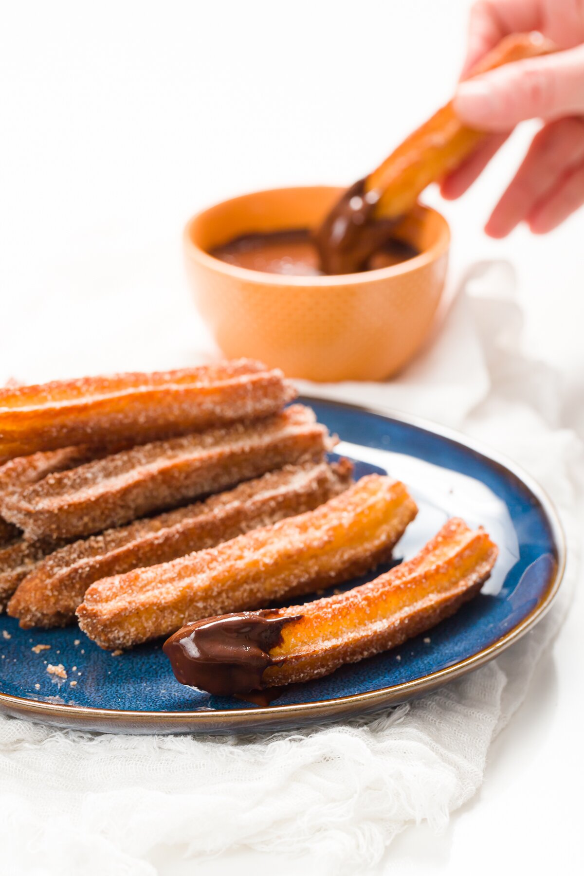 Blue plate with a stack of churros and a churros being dipped into chocolate sauce in the background