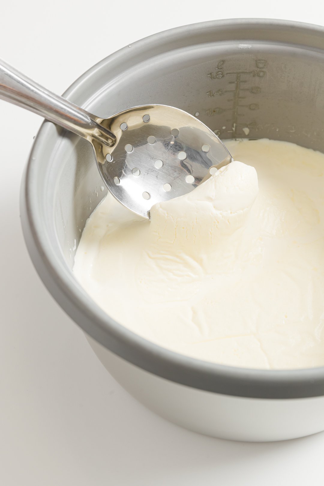 A slotted spoon skimming the clotted cream off of the top of a cream-filled rice cooker bowl