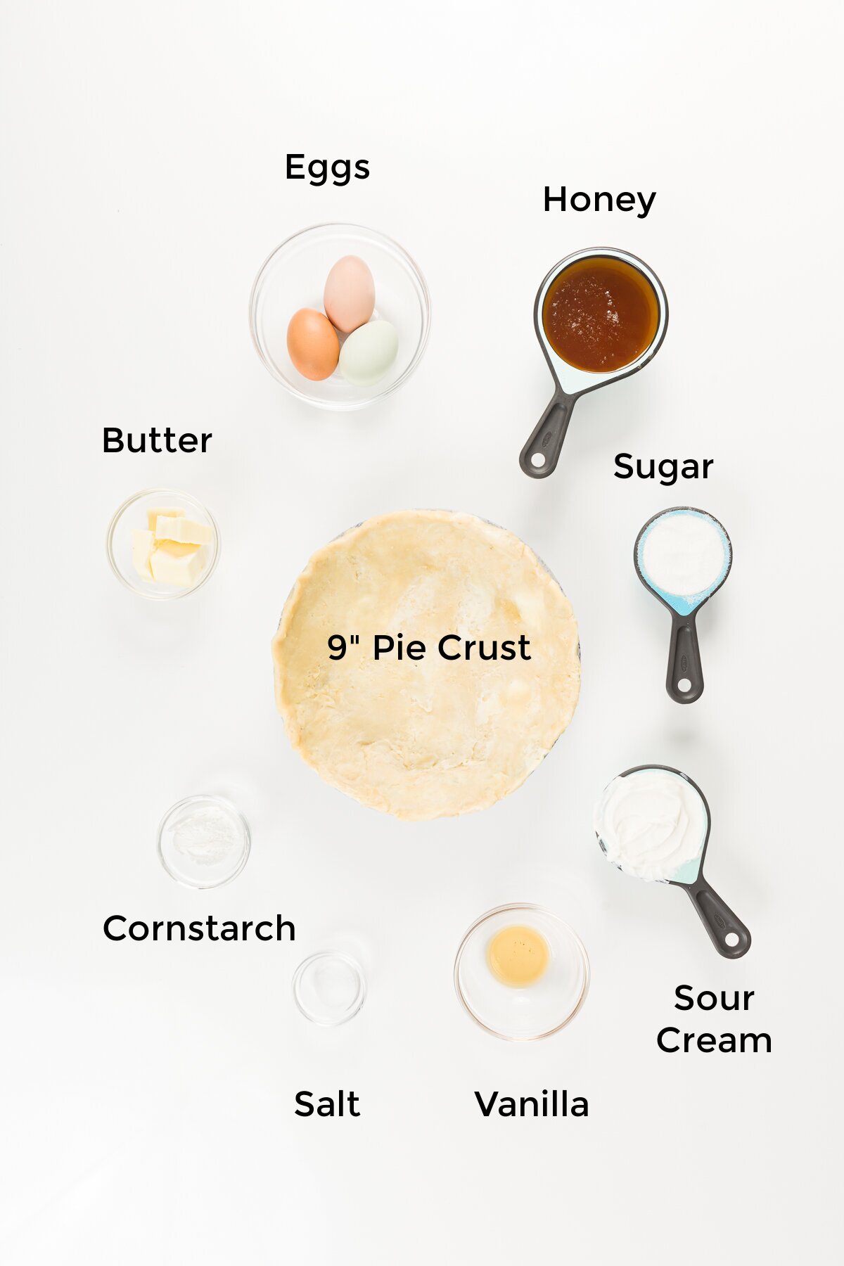 overhead view of ingredients in this honey pie recipe - eggs, honey, sugar, sour cream, vanilla extract, salt, cornstarch, butter, and a 9-inch pie crust