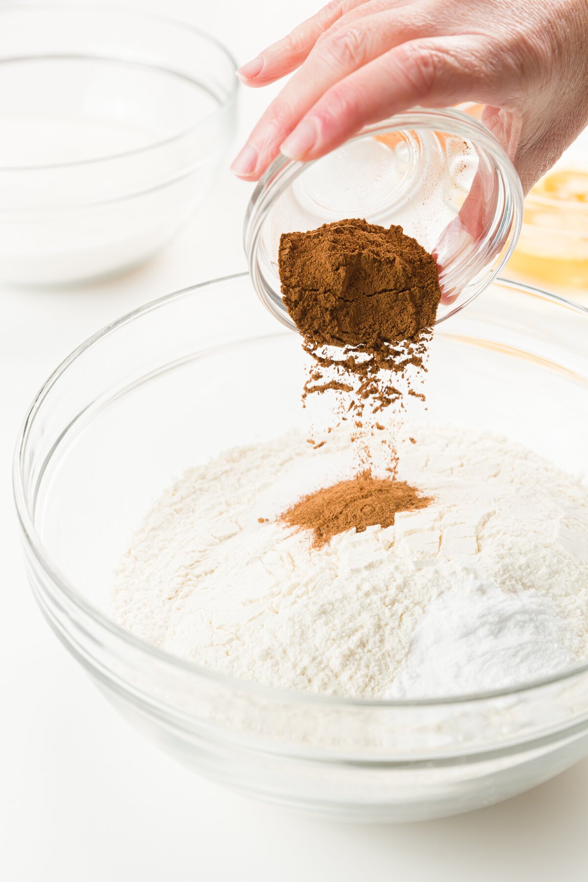 Pouring pumpkin pie spice out of a small glass blown into a larger glass bowl with dry ingredients
