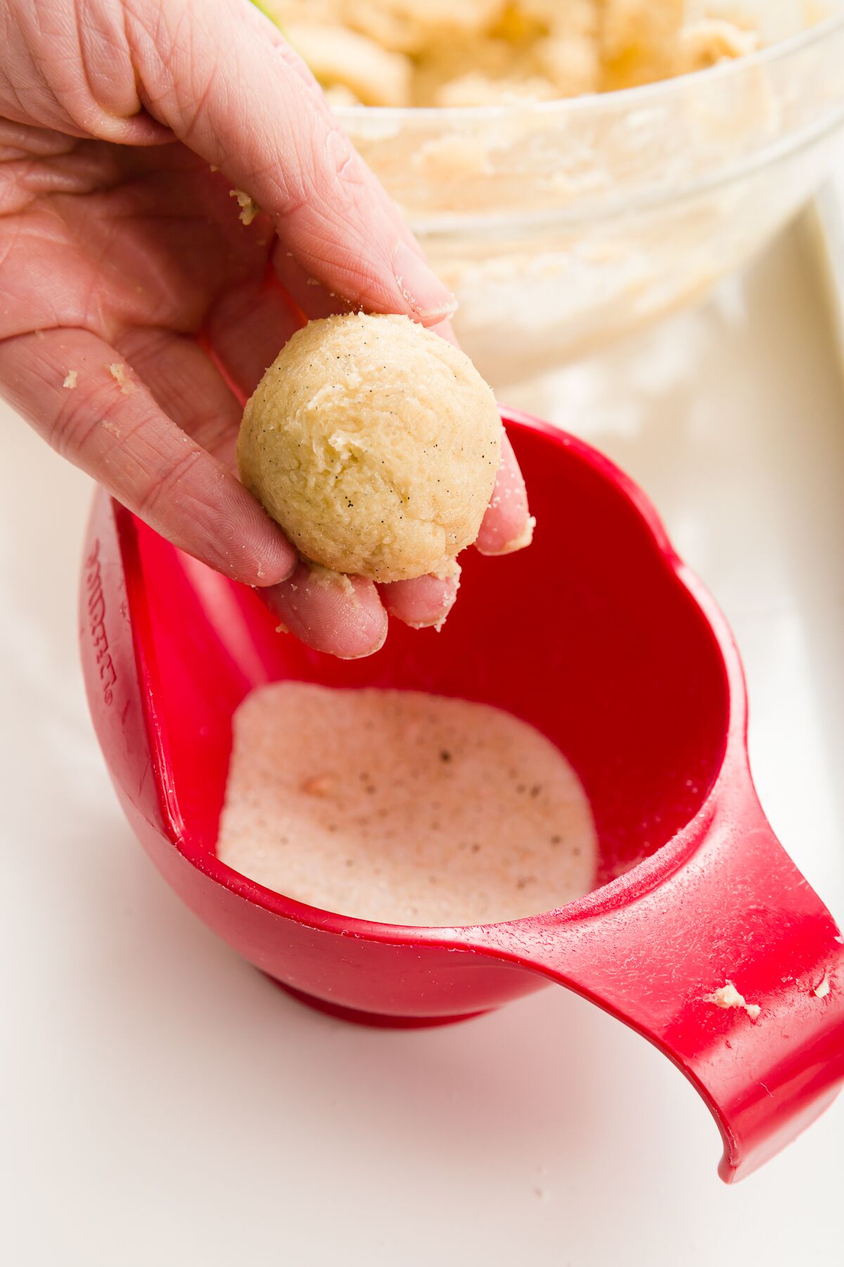 Holding a cookie dough ball over a red bowl of vanilla sugar