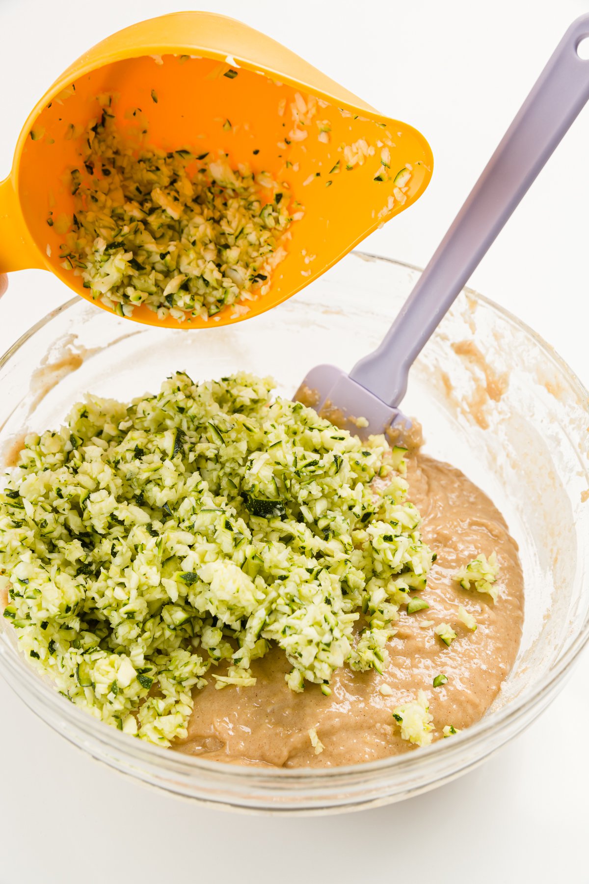Pour zucchini into bowl with batter and a purple spatula